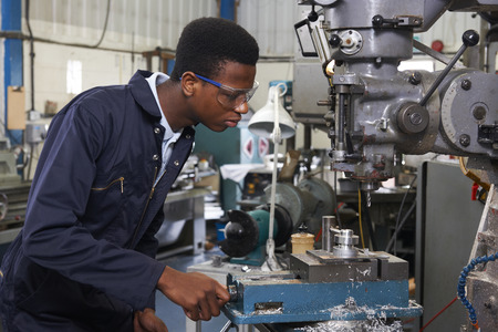 Male Apprentice Engineer Working On Drill In Factory Male Apprentice Engineer Working On Drill In Factory