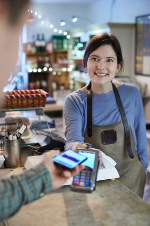 Male Customer Making Contactless Payment For Shopping Using Mobile Phone In Delicatessen の写真素材
