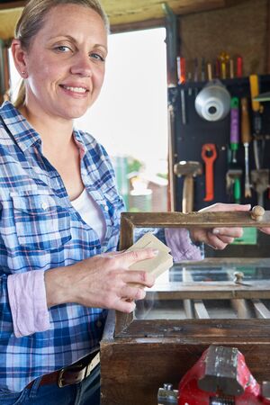 Portrait Of Mature Woman Upcycling Furniture In Workshop At Home Rubbing Down Cabinet With Sandpaperの写真素材