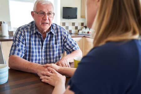 Woman Talking With Unhappy Senior Man Sitting In Kitchen At Homeの写真素材