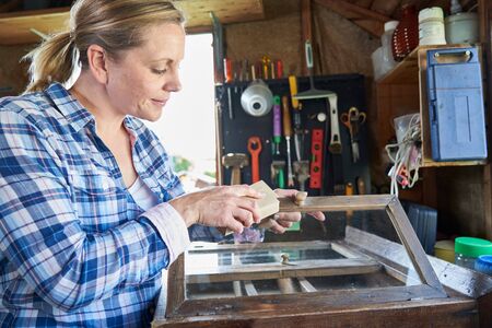 Mature Woman Upcycling Furniture In Workshop At Home Rubbing Down Cabinet With Sandpaperの写真素材