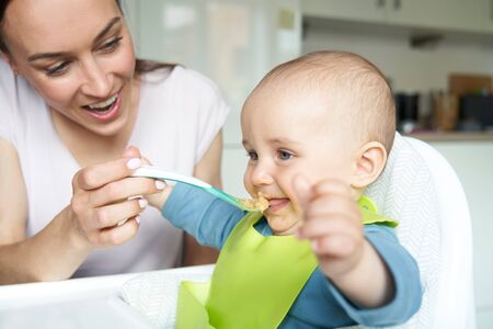 Smiling 8 month Old Baby Boy At Home In High Chair Being Fed Solid Food By Mother With Spoonの写真素材