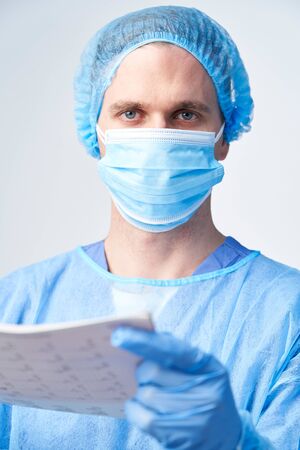 Studio Portrait Of Male Surgeon Wearing Gown And Mask Holding Medical Print Outの写真素材