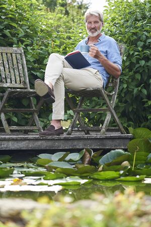 Portrait Of Mature Man Relaxing In Garden Reading Book On Jetty By Lakeの写真素材