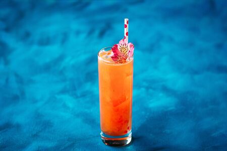 Sweet appetizing alcohol fruit cocktail with flower garnish and straw in a highball on the blue...