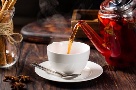 Pouring hot fresh sweet berry tea in a cup from glass teapot on the wooden table, horizontalの写真素材