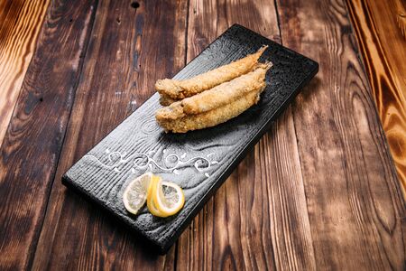 Side view on delicious deep-fried capelin with slices of lemon in a black plate on the wooden table,...