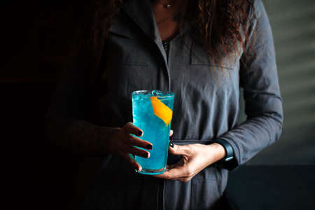 Woman holding blue cocktail in a highball with ice and orange peel, horizontal