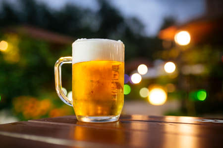 Fresh cool light beer with foam on the wooden table outside with blurry lights background, horizontalの写真素材
