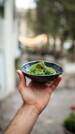 Holding black bowl with green chuka seaweed salad outside with a blurry backgroundの写真素材