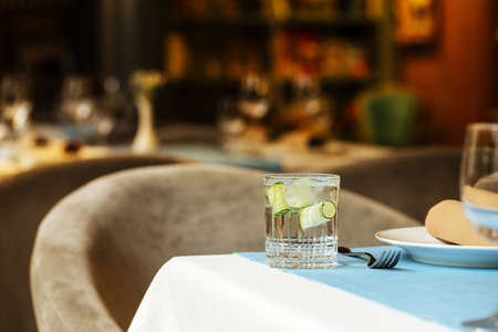 Glass of beverage with ice and cucumber on the restaurant table with a blurry lights background