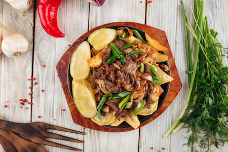 Top view on fried potatoes with beef and green onions in cast iron pan on the light wooden table