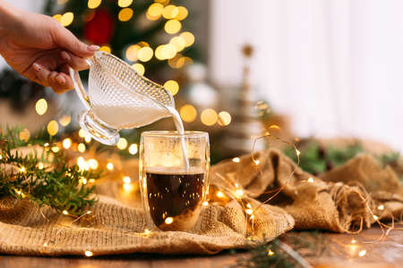 Side view on pouring milk in a glass mug of coffee on the festive garland background