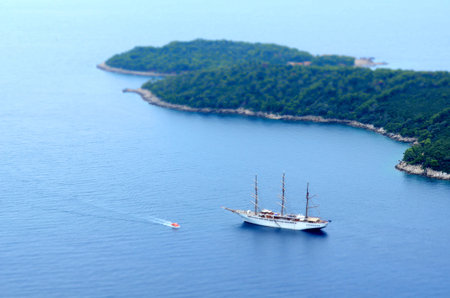 Aerial view of cruise ship and tall sailing vessel anchored near green islands in calm, clear sea, with rocky cliff in foregroundの写真素材
