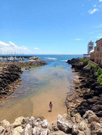 Rocky coastal channel in Cascais Portugal leading to the Atlantic Ocean with visible lighthouse and waterfront buildings under blue skyの写真素材