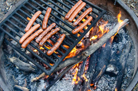 Roasting hotdog over open flame campfire. Grilling delicious meat hot dog over fire pit. Camping and cooking picnic lunch or dinner outdoors.の写真素材