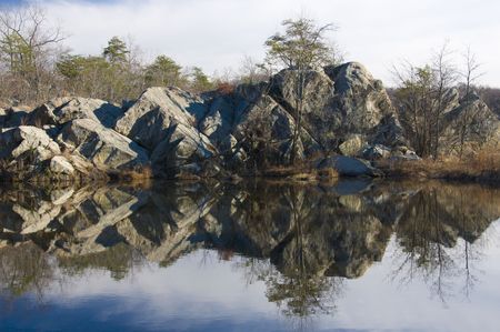 Winter Pond with steep rocky embankment and water reflections of rocks, trees, deep blue sky, and thick white cloudsの写真素材