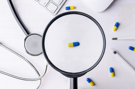 Close-up of magnifying glass and medicine with stethoscope, keyboard and mouse. Top view of white wooden working table.の写真素材