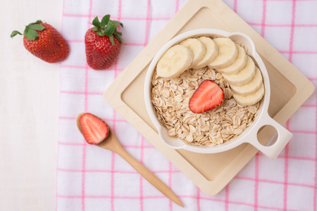 Fresh breakfast of healthy oatmeal with banana slices, strawberries, blueberries and milk in a bowl on white wooden backgroundの写真素材