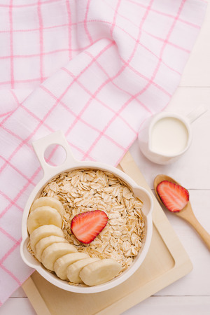 Fresh breakfast of healthy oatmeal with banana slices, strawberries, blueberries and milk in a bowl on white wooden backgroundの写真素材