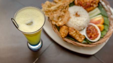 set of menu of a glass of juice and tradisional indonesian cuisine; ayam penyet, fried chicken served with white rice, chili sauce,slices of cucumber, tomato, and chips served on bamboo plaited plateの写真素材