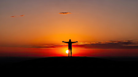 A dark silhouette of a person stands with arms open, facing a vivid orange and red sunset. The composition features a low angle with a clear sky and the sun at the horizon. This image conveys a sense of freedom and could be suitable for various commercial or editorial uses related to travel or nature.の素材