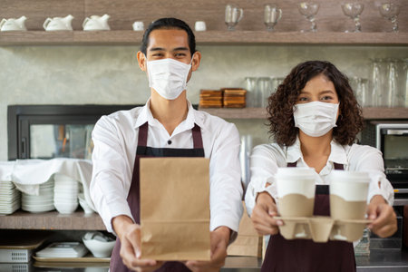 Asian barista with mask holding paper bag and coffee cup for customer take away for provent coronavirus in restaurant.の写真素材