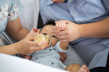 Asian baby drink milk.Thai parent holding little child hands for feed food in living room.の写真素材