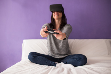 Casual young woman sitting on the bed at home having fun playing a virtual reality video gameの写真素材