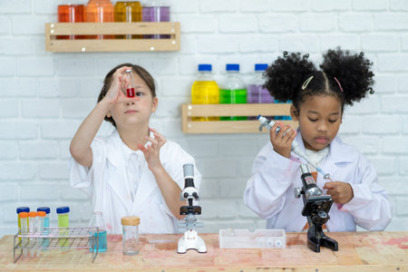 Two little kids in lab coat learning chemistry in school laboratory. Young scientists in protective glasses making experiment in lab or chemical cabinet. Working on a tablet.の写真素材
