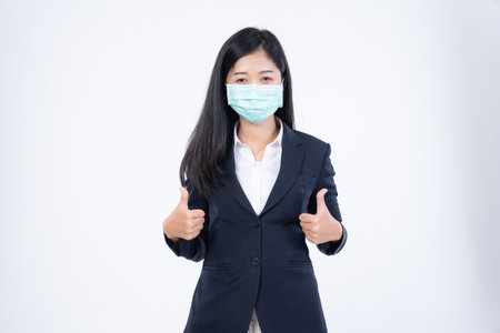 A confident beautiful young Asian business woman wearing green medical face mask and black suit, standing with her arms folded and looking at camera isolated on white background.の写真素材