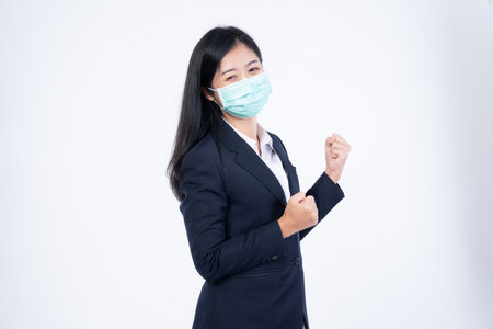 A confident beautiful young Asian business woman wearing green medical face mask and black suit, standing with her arms folded and looking at camera isolated on white background.の写真素材