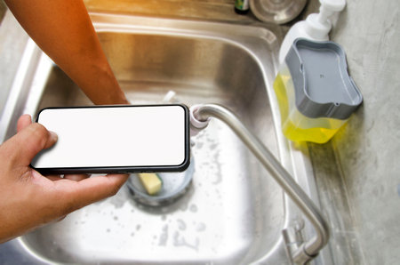 Right hand holding blank screen smartphone or mobile phone while washing dish in sink of kitchen.using technology for communication while housework and working concept.View of copy space.の写真素材