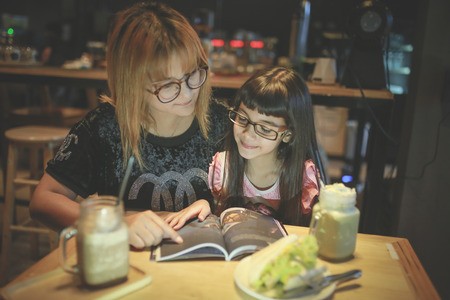 The mother of an thai Asian child with glasses is about to read a book in a coffee shop.の写真素材