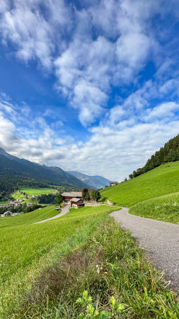 Santa Maddalena, Val di Funes,Quaint house, winding road, lush pastures, clear blue sky with fluffy clouds, framed by layered Dolomites-an Alpine serenityの写真素材
