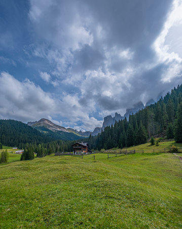 Nestled in Val di Funes, a wooden cabin stands against the Dolomites backdrop, surrounded by lush green pastures, a serene alpine retreat embraced by natureの写真素材