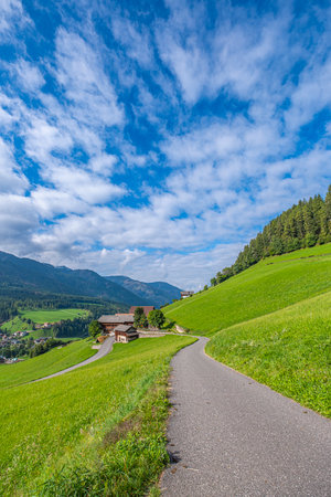Santa Maddalena, Val di Funes, A quaint house along a winding road, embraced by lush pastures, guides to the majestic, layered Dolomites, an enchanting Alpine spectacleの写真素材