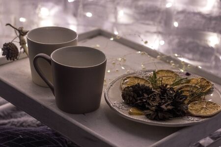 two white mugs on a wooden tray stand on a bed with a fluffy blanket with a garland in the backgroundの写真素材