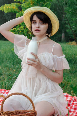 Beautiful romantic young woman with dark short hair in a straw hat and white dress is sitting on a picnic, in nature with a bottle of milkの写真素材