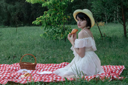 Beautiful romantic young woman with dark short hair in a straw hat and white dress is sitting on a picnic, in natureの写真素材