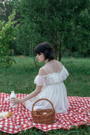 Beautiful romantic young woman with dark short hair in a straw hat and white dress is sitting on a picnic, in nature with a bottle of milkの写真素材