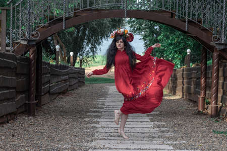 beautiful young girl in a red dress with long sleeves and a wreath is dancing under the bridge in the parkの写真素材