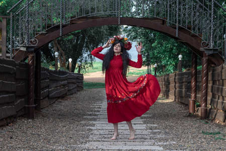 beautiful young girl in a red dress with long sleeves and a wreath is dancing under the bridge in the parkの写真素材