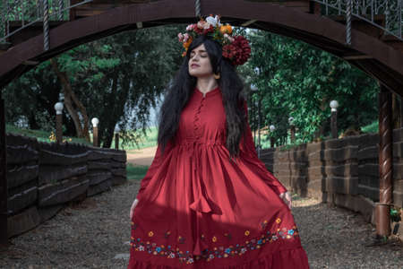 beautiful young girl in a red dress with long sleeves and a wreath is dancing under the bridge in the parkの写真素材