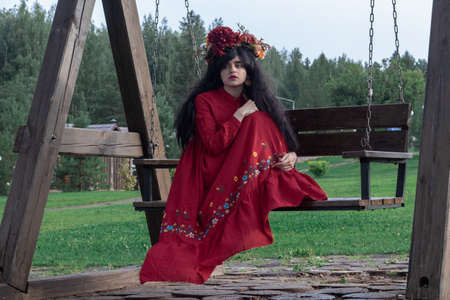 beautiful young girl in a red dress with long sleeves and a wreath sits on a wooden swingの写真素材