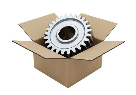 A close-up studio shot of a single, shiny metal gear with prominent teeth, positioned inside an open, light brown cardboard box. The background is pure white.の素材