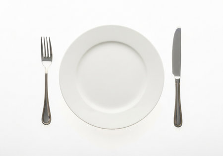 An overhead view captures a clean, white ceramic plate centered between a gleaming silver fork and knife, all set neatly on a plain white background, ready for a meal.の素材