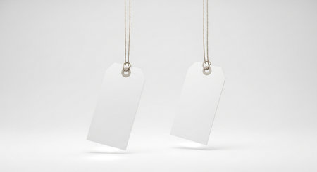 Two blank white price tags are suspended by strings against a clean, neutral white background, offering ample space for customization and branding.の素材
