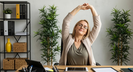 A woman sits at her office desk, wearing a light blazer and glasses, stretching her arms overhead with eyes closed, finding a moment of relaxation and rejuvenation.の素材
