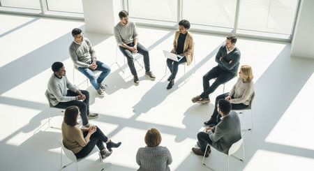 A diverse group of individuals are seated in a circle, engaged in a discussion, promoting a sense of community and shared experience in a well-lit, open space.の素材